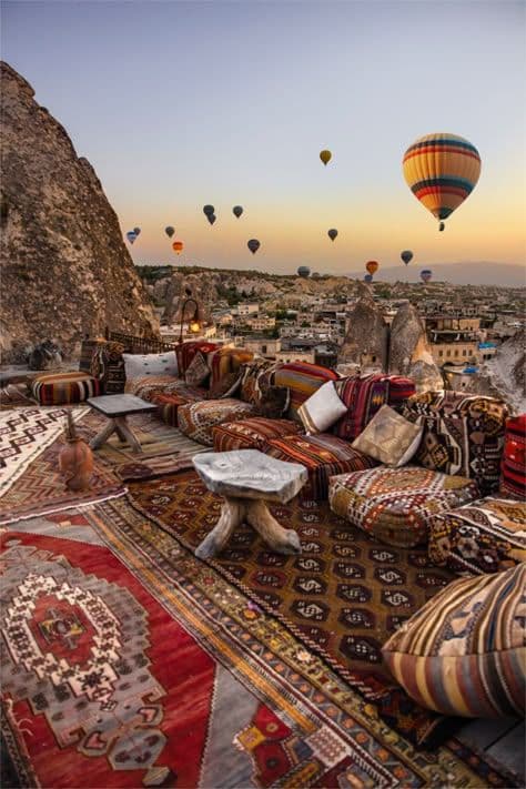 Sunrise hot air balloons over a cliffside terrace with patterned rugs and cushions.